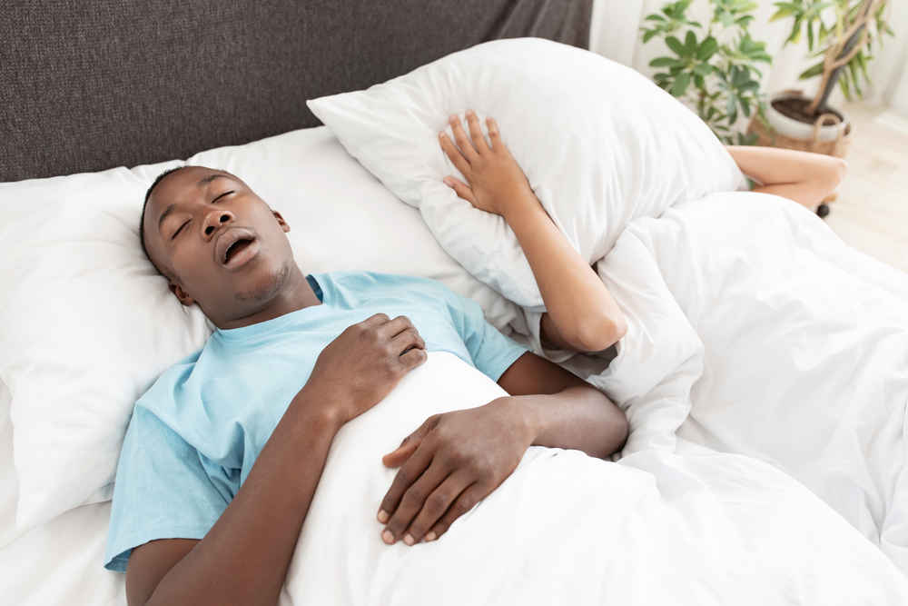 Man snoring due to sleep apnea while wife is covering her ears and face with a pillow in bed
