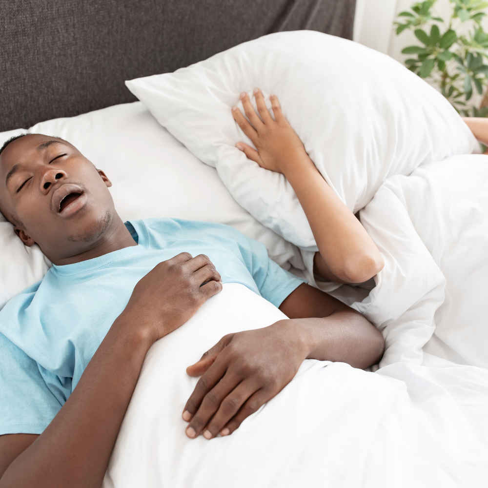 Man snoring due to sleep apnea while wife is covering her ears and face with a pillow in bed